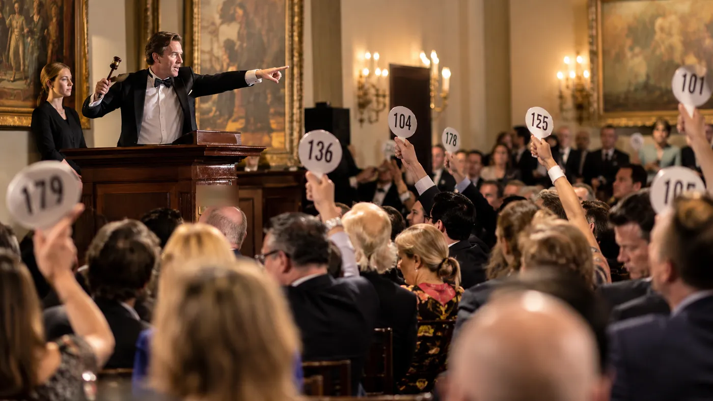 An auctioneer raising a gavel as bidders lift numbered paddles in an auction room (illustration)
